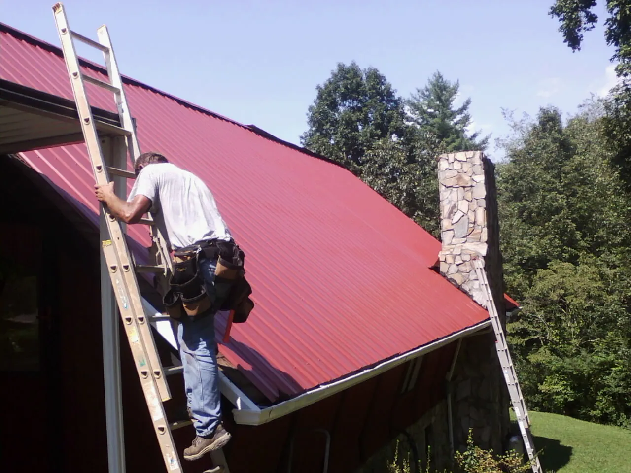 Roofer on a ladder beside a red metal roof and stone chimney — Soffit & Fascia Repair by Roofing Brothers in Advance